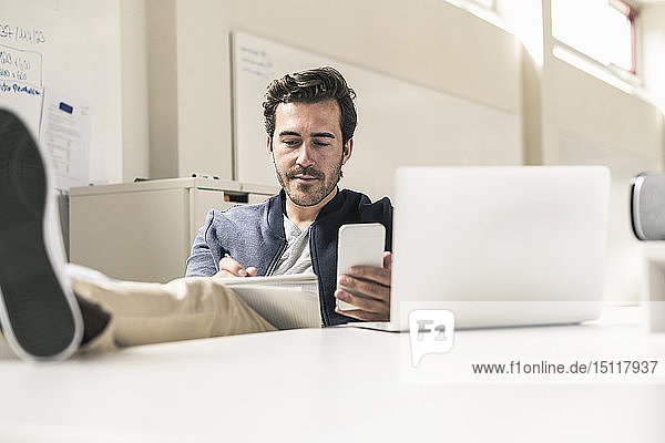 Young businessman working relaxed in modern office using smartphone and laptop