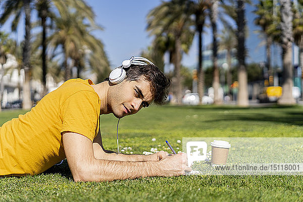 Spain  Barcelona  man lying on lawn in the city with headphones and notebook