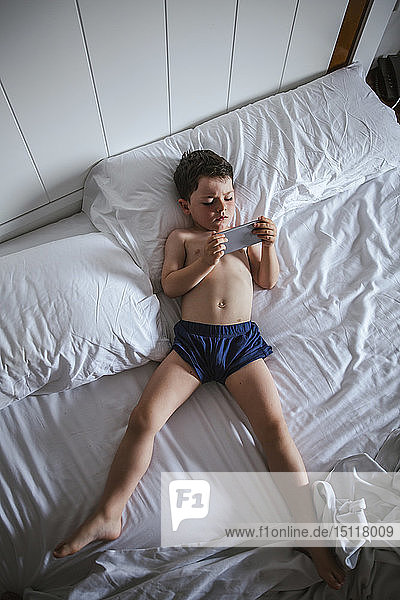 Kleiner Junge liegt auf dem Bett und benutzt ein Smartphone