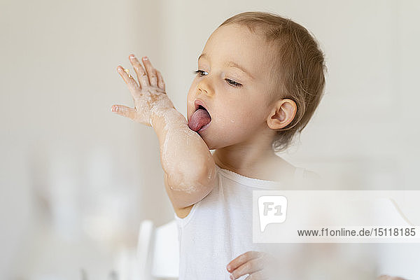 Little girl licking her arm while making a cake in kitchen at home