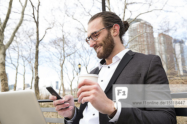 Smiling young businessman with coffee to go looking at cell phone  New York City  USA