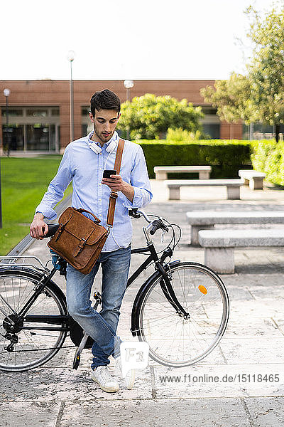 Young man with bicycle using smartphone  headphones around neck in the city