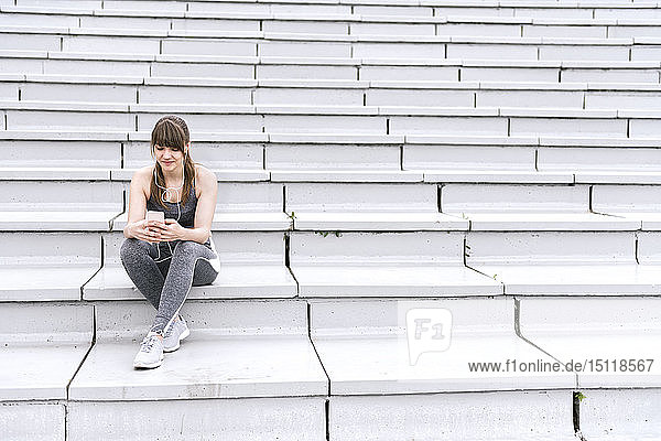 Junge Frau in Sportkleidung sitzt auf einer Betontribüne und hört Musik auf einem Smartphone