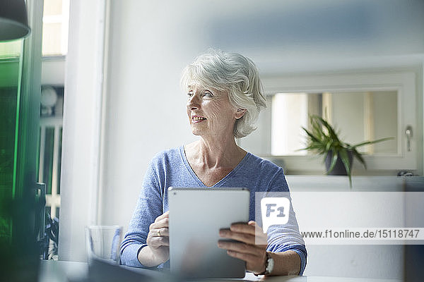 Lächelnde reife Frau mit digitalem Tablett  die aus dem Fenster schaut