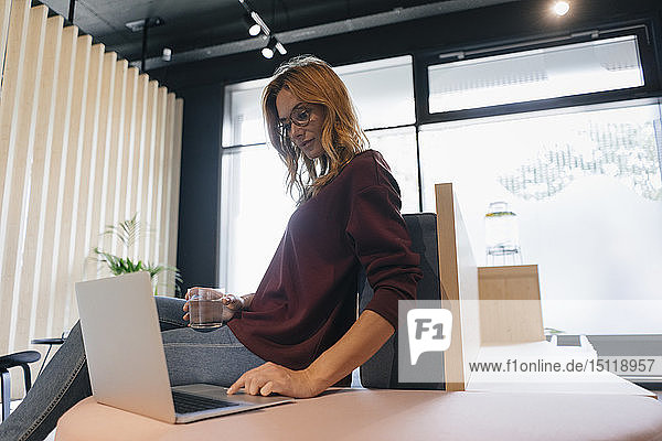 Businesswoman sitting on desk in office using laptop