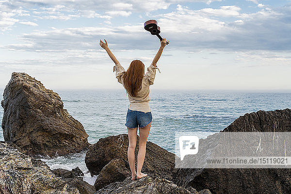 Rückenansicht einer jungen Frau mit Ukulele  die auf einem Felsen steht und auf das Meer schaut
