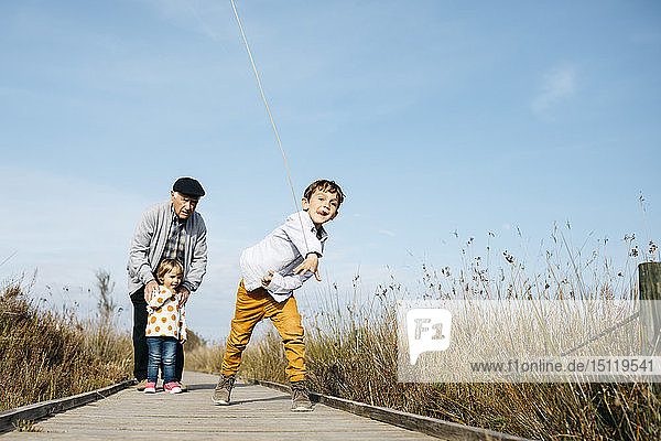 Junge auf der Strandpromenade beim Stockwerfen  während sein Großvater und seine kleine Schwester ihn aus dem Hintergrund beobachten