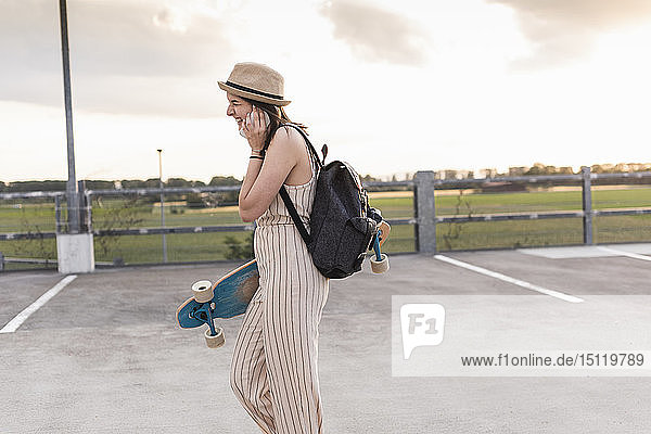 Happy young woman with cell phone and longboard walking on parking deck
