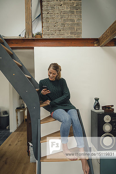 Lächelnde junge Frau sitzt zu Hause auf der Treppe und benutzt ihr Handy