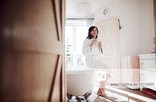 Reife Frau sitzt auf Badewanne und trinkt Kaffee