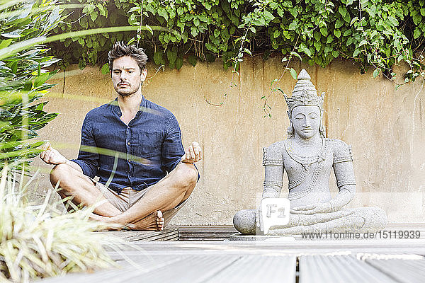 Mann sitzt im Schneidersitz neben der Buddhastatue in einem Zen-Garten und meditiert