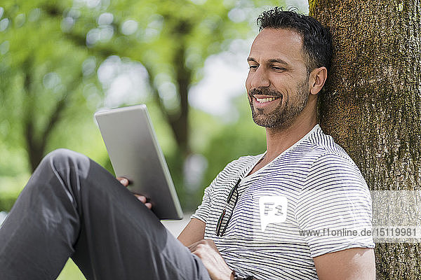 Lächelnder Mann lehnt mit Tablette an einen Baum im Park