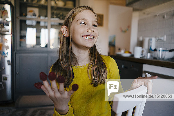 Happy girl sitting at kitchen table at home playing with raspberries