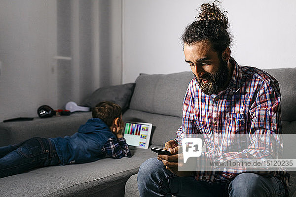Vater sitzt auf der Couch und schaut auf ein Smartphone  während sein Sohn im Hintergrund ein digitales Tablet benutzt