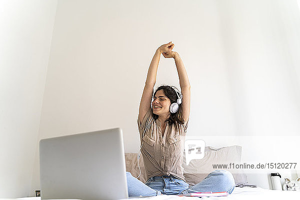 Glückliche junge Frau sitzt mit Kopfhörern und Laptop auf dem Bett