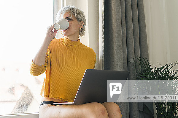 Frau sitzt zu Hause am Fenster mit Laptop und trinkt Kaffee aus Tasse