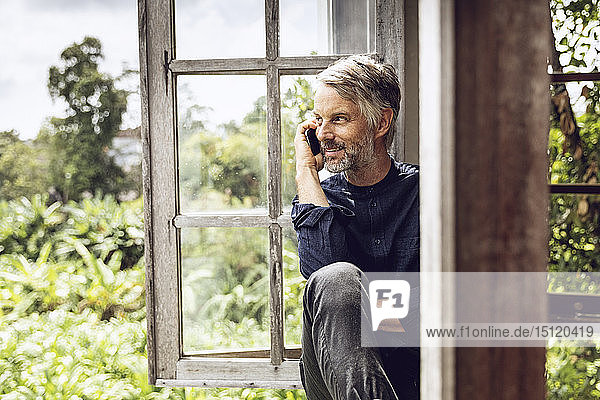 Mann am Handy sitzt am Fenster in tropischer Umgebung
