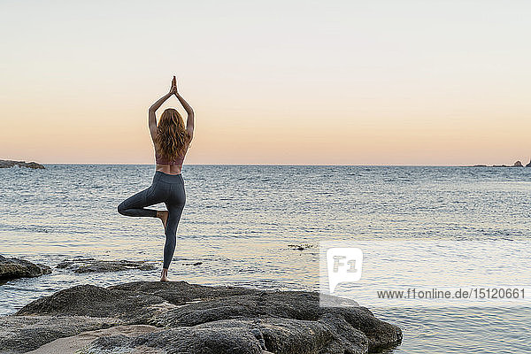 Junge Frau praktiziert Yoga am Strand  in Baumpose  während des Sonnenuntergangs am ruhigen Strand  Costa Brava  Spanien