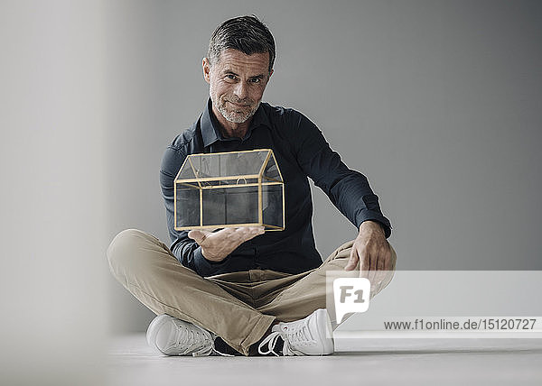 Confident mature businessman sitting on the floor holding glass house model
