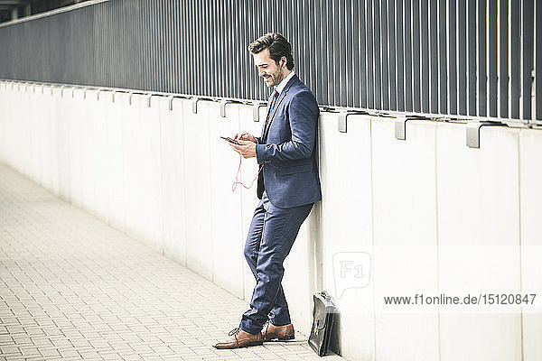 Businessman leaning against a wall in the city checking cell phone
