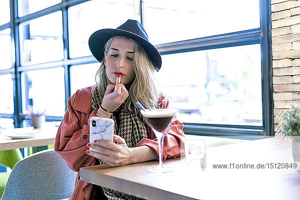Woman holding cell phone applying lipstick in a cafe