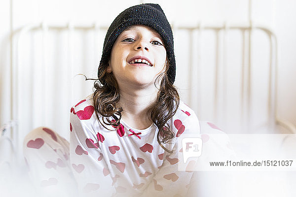 Porträt eines lächelnden kleinen Mädchens  das mit Mütze und Pyjama im Bett sitzt