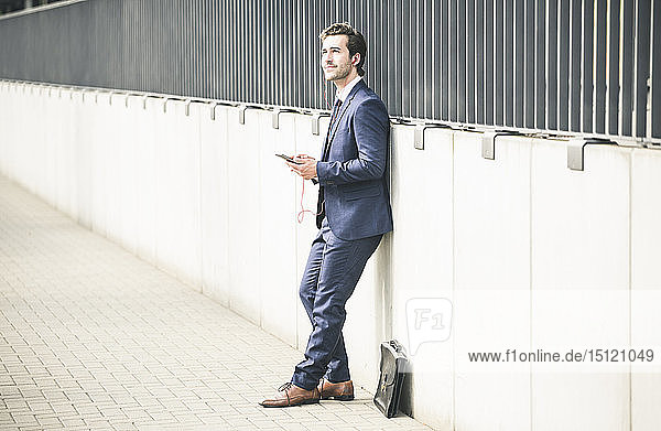 Businessman leaning against a wall in the city with cell phone and earphones