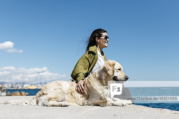 Glückliche Frau sitzt neben ihrem Labrador Retriever am Kai und schaut auf das Meer