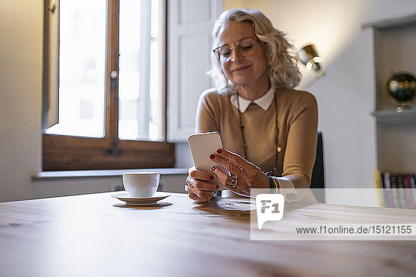 Reife Geschäftsfrau  die mit einem Smartphone bei einer Tasse Kaffee am Tisch sitzt