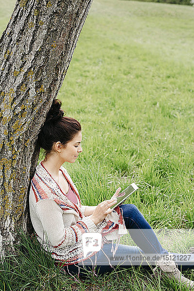 Junge Frau sitzt mit Tablette an einem Baum auf dem Land