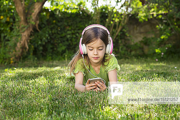 Portrait of little girl lying on meadow listening music with headphones using smartphone
