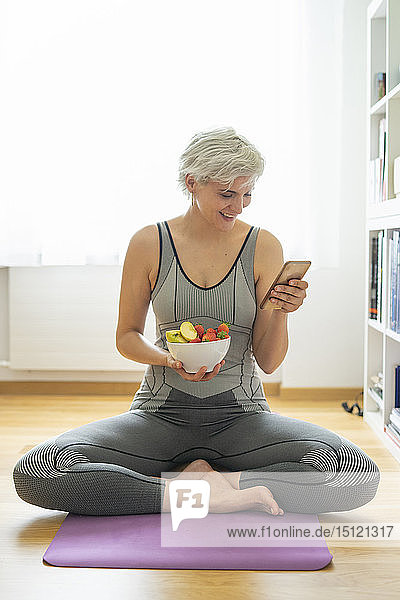 Frau  die zu Hause Yoga praktiziert  einen gesunden Fruchtsnack zu sich nimmt und ein Smartphone benutzt