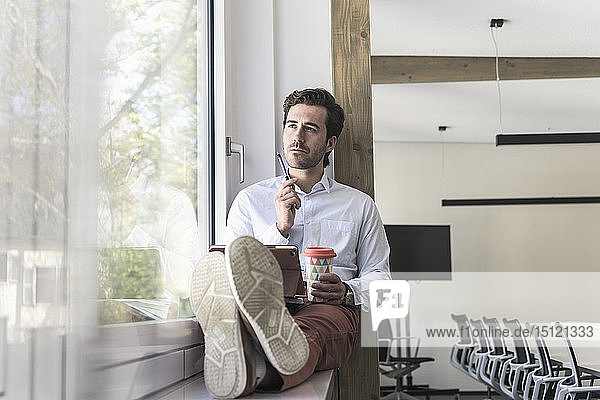 Young businessman sitting on windowsill  using digital tablet  drinking coffee