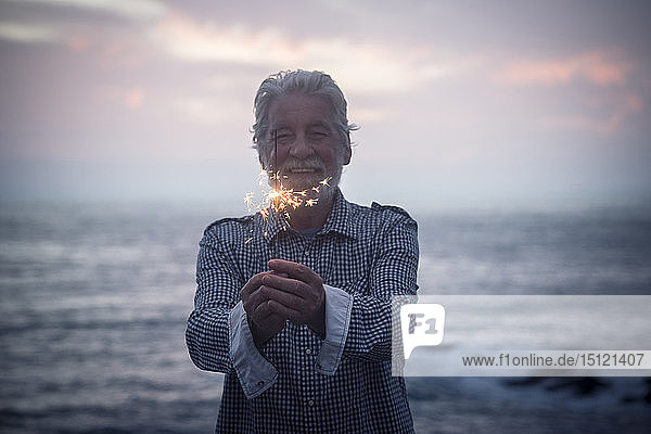 Glücklicher älterer Mann steht bei Sonnenuntergang mit einer Wunderkerze vor dem Meer