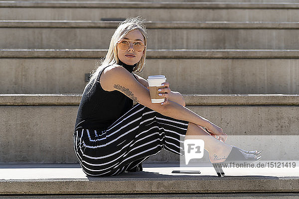 Bortrait of blond young woman with coffee to go sitting on stairs outdoors