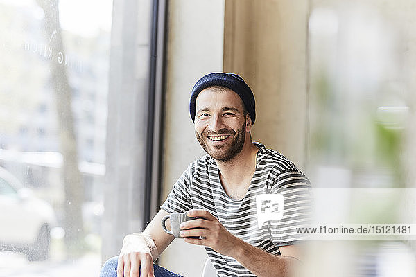 Porträt eines lächelnden jungen Mannes  der eine Kaffeetasse am Fenster hält