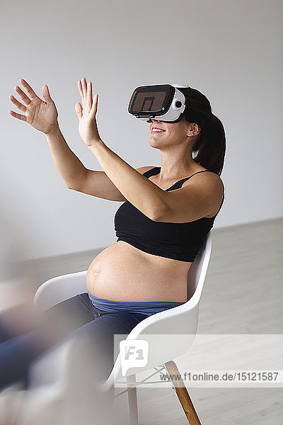 Junge schwangere Frau  die in einem Sessel sitzt und ihr Baby mit Hilfe einer Virtual-Reality-Brille beobachtet und ertastet