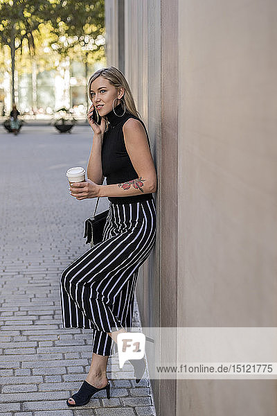 Portrait of smiling blond woman with coffee to go on the phone leaning against wall