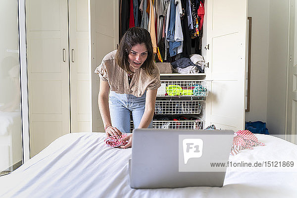 Junge Frau mit Laptop legt Kleider aufs Bett