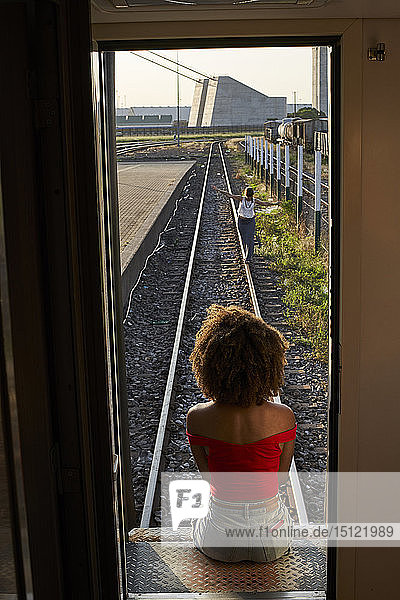 Frau  die an der Hintertür eines Zuges sitzt und eine Frau auf den Gleisen ansieht