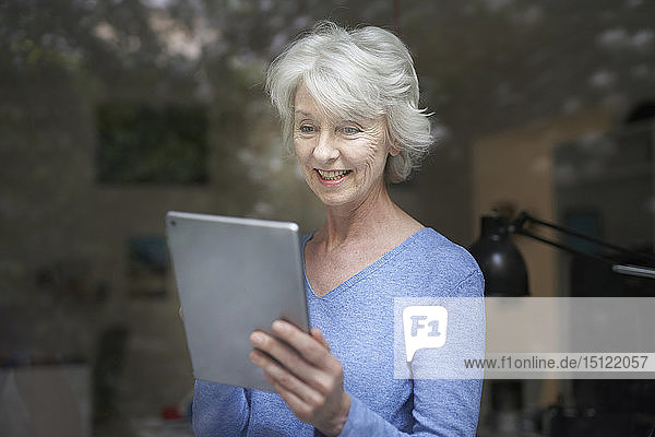 Portrait of smiling mature woman behind windowpane looking at digital tablet