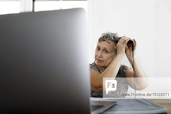 Ältere Frau sitzt zu Hause am Tisch mit Laptop und macht sich die Haare