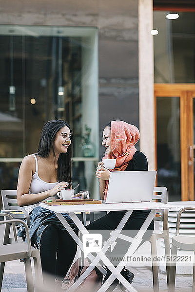 Zwei glückliche Freunde sitzen mit Laptop in einem Straßencafé zusammen