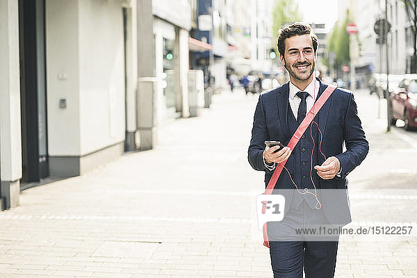 Lächelnder Geschäftsmann  der mit Handy und Kopfhörern durch die Stadt läuft