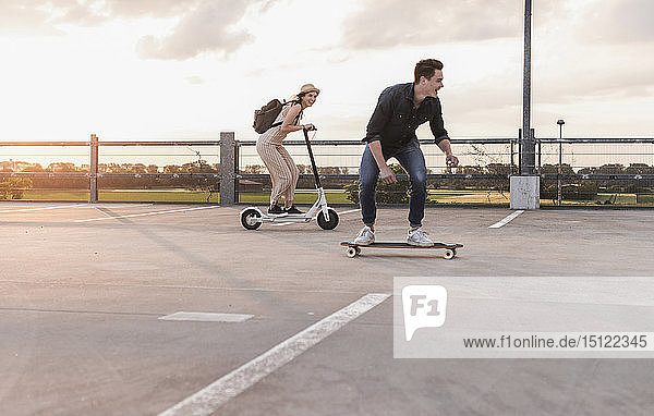 Young man and woman riding on longboard and electric scooter on parking deck at sunset