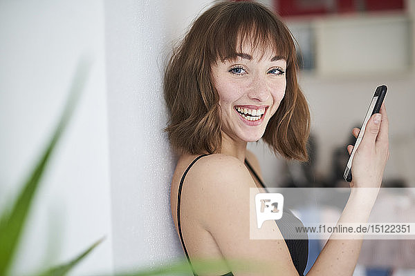 Porträt einer lachenden jungen Frau mit Smartphone zu Hause