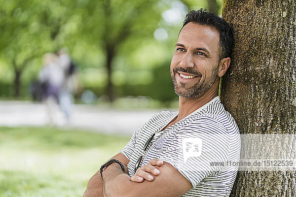Porträt eines lächelnden Mannes  der sich im Park an einen Baum lehnt