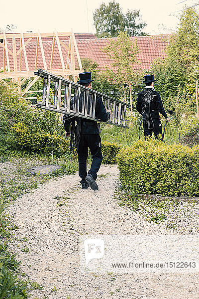 Zwei Schornsteinfeger gehen mit Leiter auf dem Gartenweg