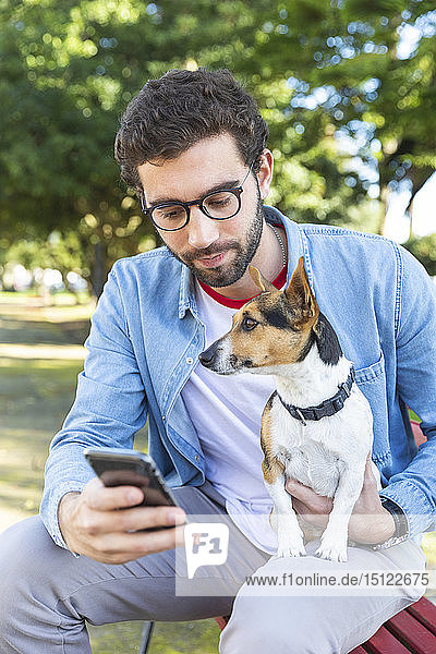 Portrait of young man sitting on park bench with his dog looking at smartphone