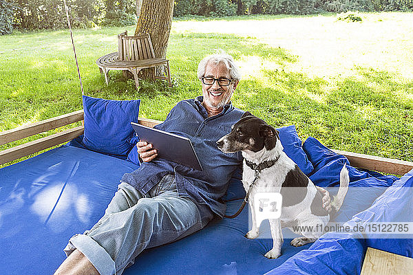 Älterer Mann entspannt sich auf einem Schaukelbett in seinem Garten und benutzt ein digitales Tablet